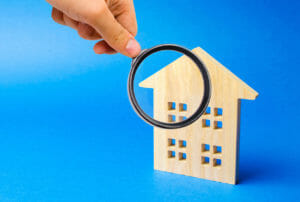 A hand holding a magnifying glass over a wooden cutout of a house.